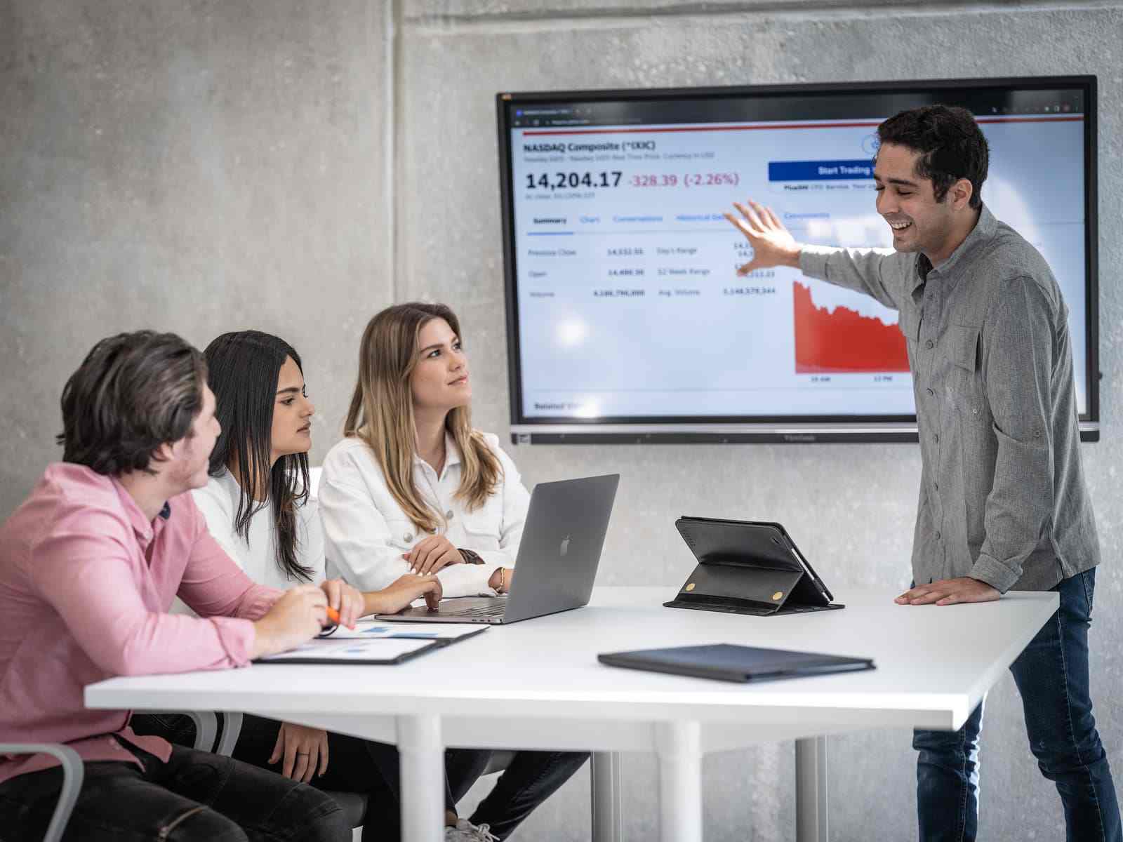 Joven señala un gráfico financiero proyectado en una pantalla mientras tres compañeros lo observan durante una presentación en sala de reuniones.